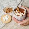 Hot chocolate with marshmallows and a cinnamon stick, held by a hand on a textured surface.