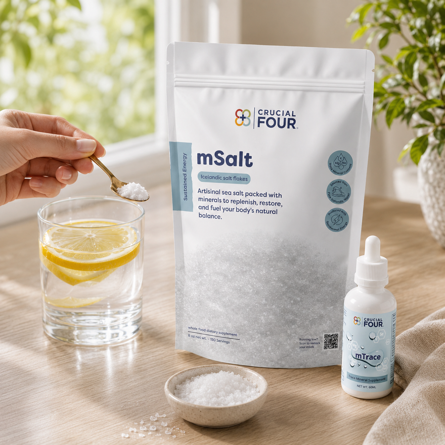 A hand holds a spoonful of white mSalt supplement powder beside a glass of water with a lemon slice, with a large white Crucial Four mSalt pouch and a small white dropper bottle on a wooden table in a bright indoor setting.