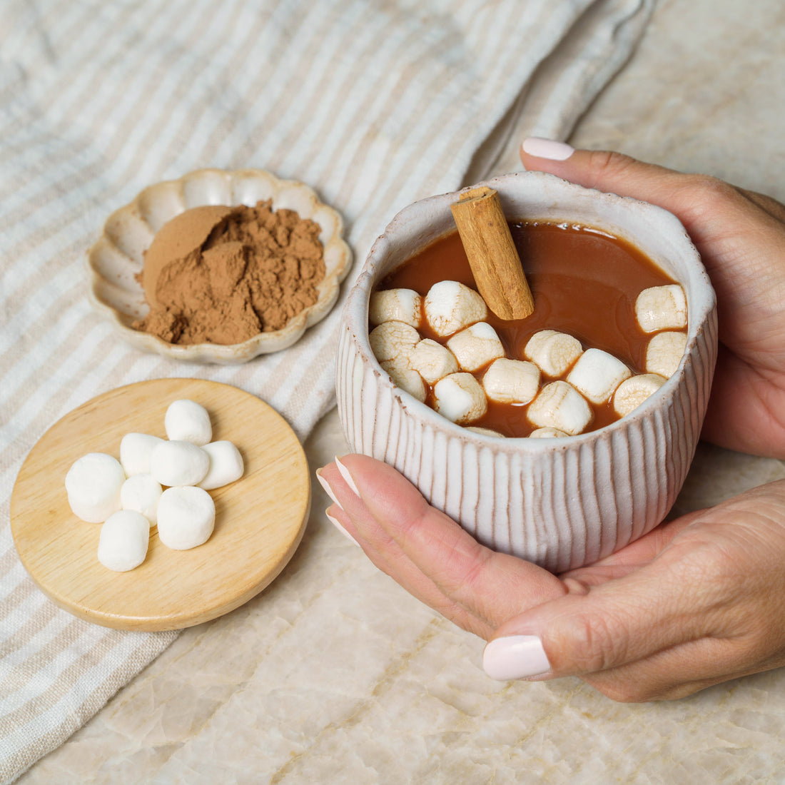 Hot chocolate with marshmallows and a cinnamon stick, held by a hand on a textured surface.